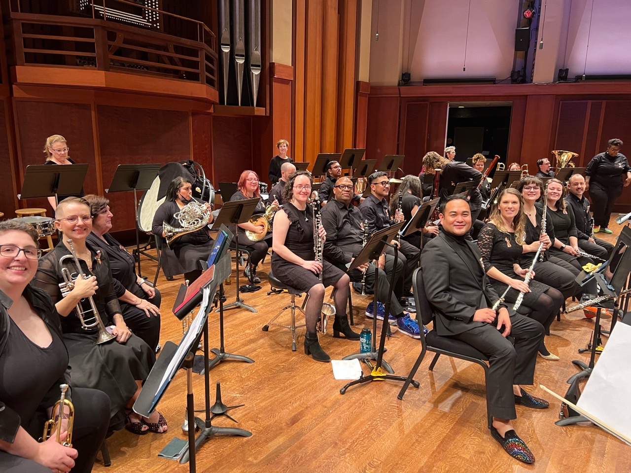 Photo of winds section in Rainbow City Orchestra with flute & bass clarinet to the right and trumpets on the left. Performers are dressed in all black seated in folding chairs placed on a wooden stage.